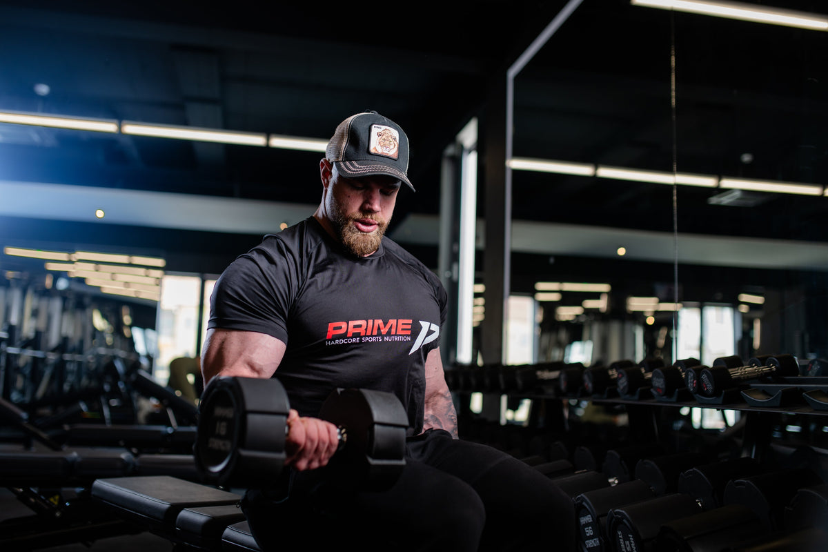 Bărbat cu barbă, șapcă și tricou negru Prime Hardcore Sports Nutrition face flexii cu gantera, așezat pe bancă