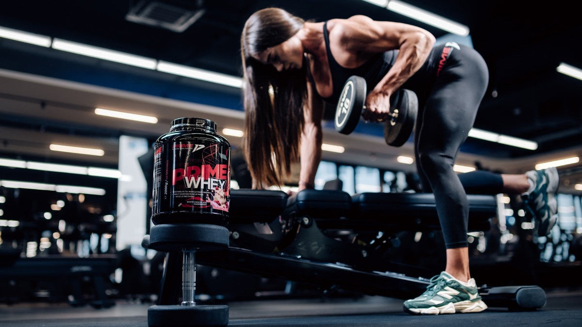 Sportivă la ramat cu gantera pe bancă, cu un tub de Prime Whey 2 kg lângă bancă, în sala de forță