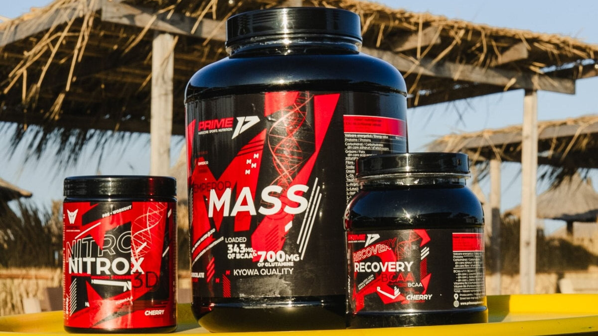 Gainerul Emperor Mass lângă Nitrox 3D și Recovery BCAA pe o masă galbenă, pe plajă, sub umbrele de stuf